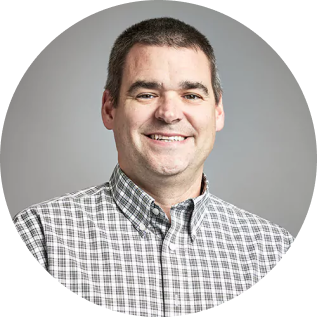 A man with short dark hair, smiling, wearing a new plaid button-down shirt, in front of a gray background.