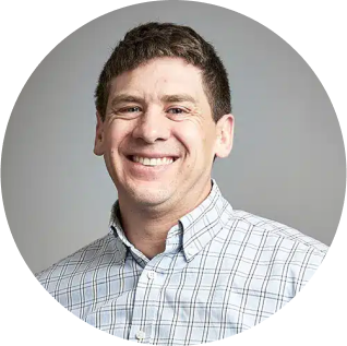 A man with short brown hair is smiling, wearing a white and blue checkered shirt. He is facing forward against a plain grey background, looking about ready for something new.