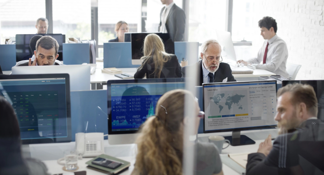 Office workers at desks with computers displaying federal financial data and maps, focused on their tasks in a busy open-plan office.