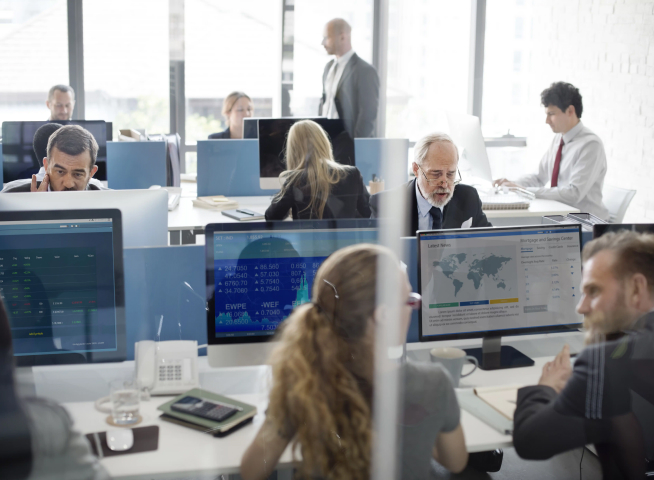 In the new office environment, multiple people are working at desks, each using computers. Various screens display data and graphs. Several professionals are engaged in conversations or focused on tasks, discussing about different projects and strategies.