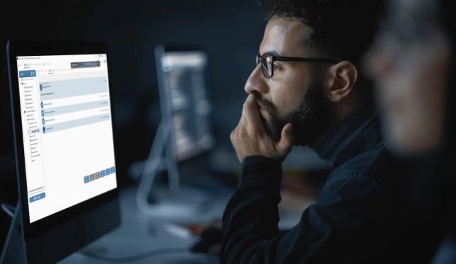 A man with glasses is sitting in a federal office, looking intently at a computer screen displaying a software interface. Another person is out of focus in the foreground.