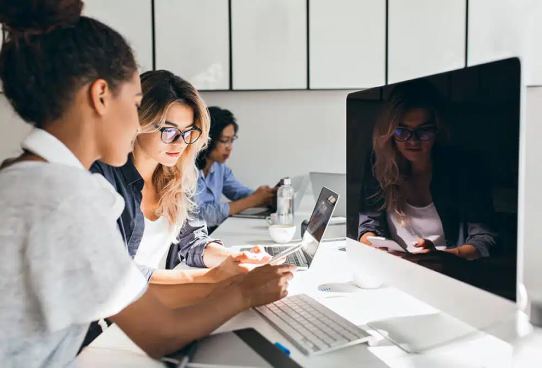 Four diverse women focused on their work in a bright office, using laptops and desktop computers for higher education IT solutions.