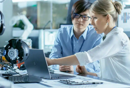 Two engineers, a man and a woman, discuss an IT solutions project on a laptop in a high-tech lab with electronic components on the table.