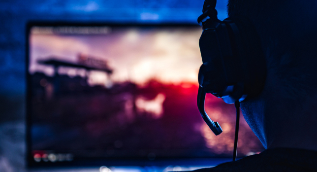 A student in higher education wears a headset, immersed in playing a video game on a screen in a dimly lit room.